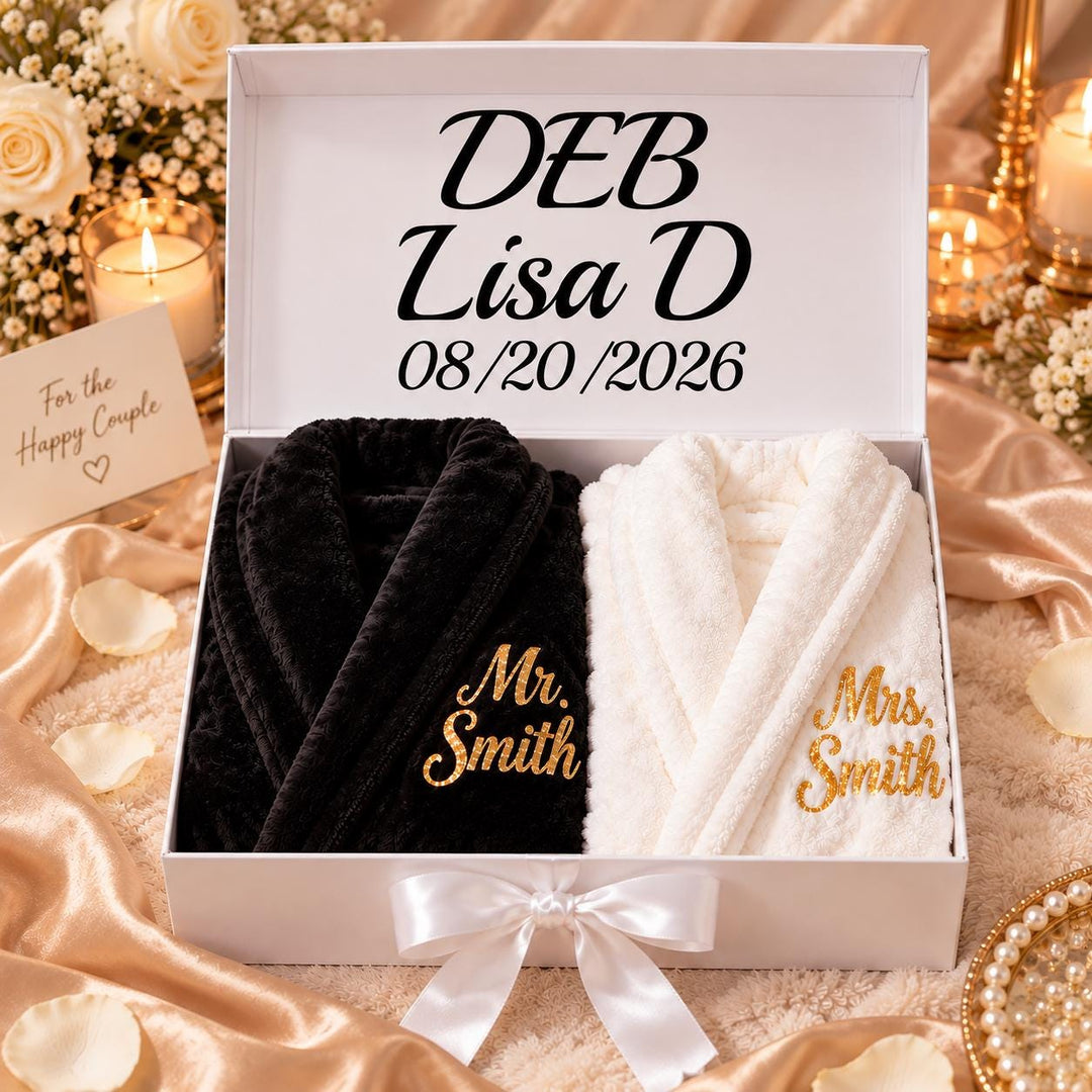 A white box containing two black and white robes with gold embroidery, placed on a table with a floral arrangement and candles.