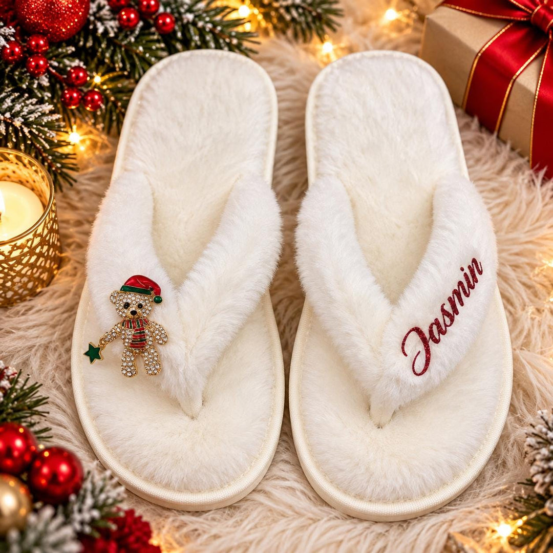 A pair of white slippers with a teddy bear charm and the name "Jasmin" embroidered on them, placed on a white furry surface surrounded by Christmas decorations and a gift box.