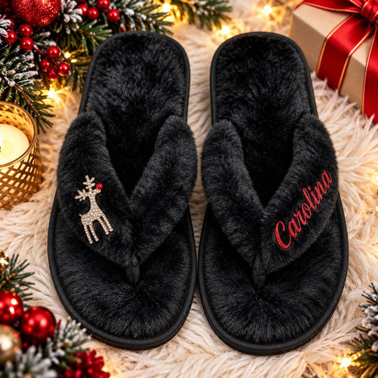 A pair of black slippers with a deer charm and the name "Carolina" embroidered on them, placed on a white surface with Christmas decorations and a gift box in the background.