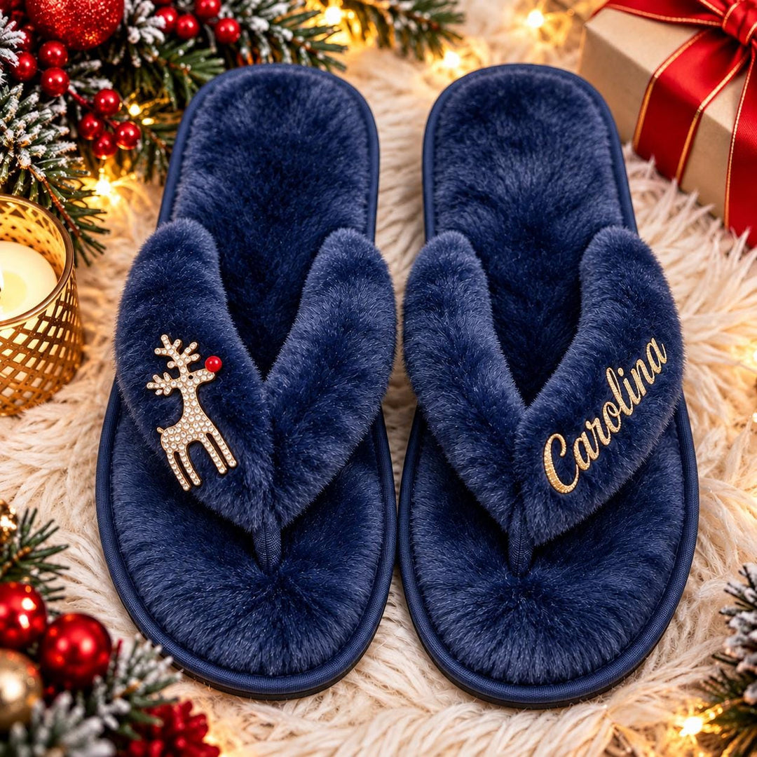 A pair of blue slippers with a reindeer charm and the name "Carolina" embroidered on them, placed on a white surface with Christmas decorations and a gift box in the background.