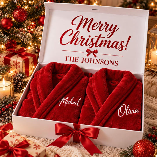 A white gift box containing two red bathrobes with personalized names on them, placed on a table with a Christmas tree and other holiday decorations in the background.