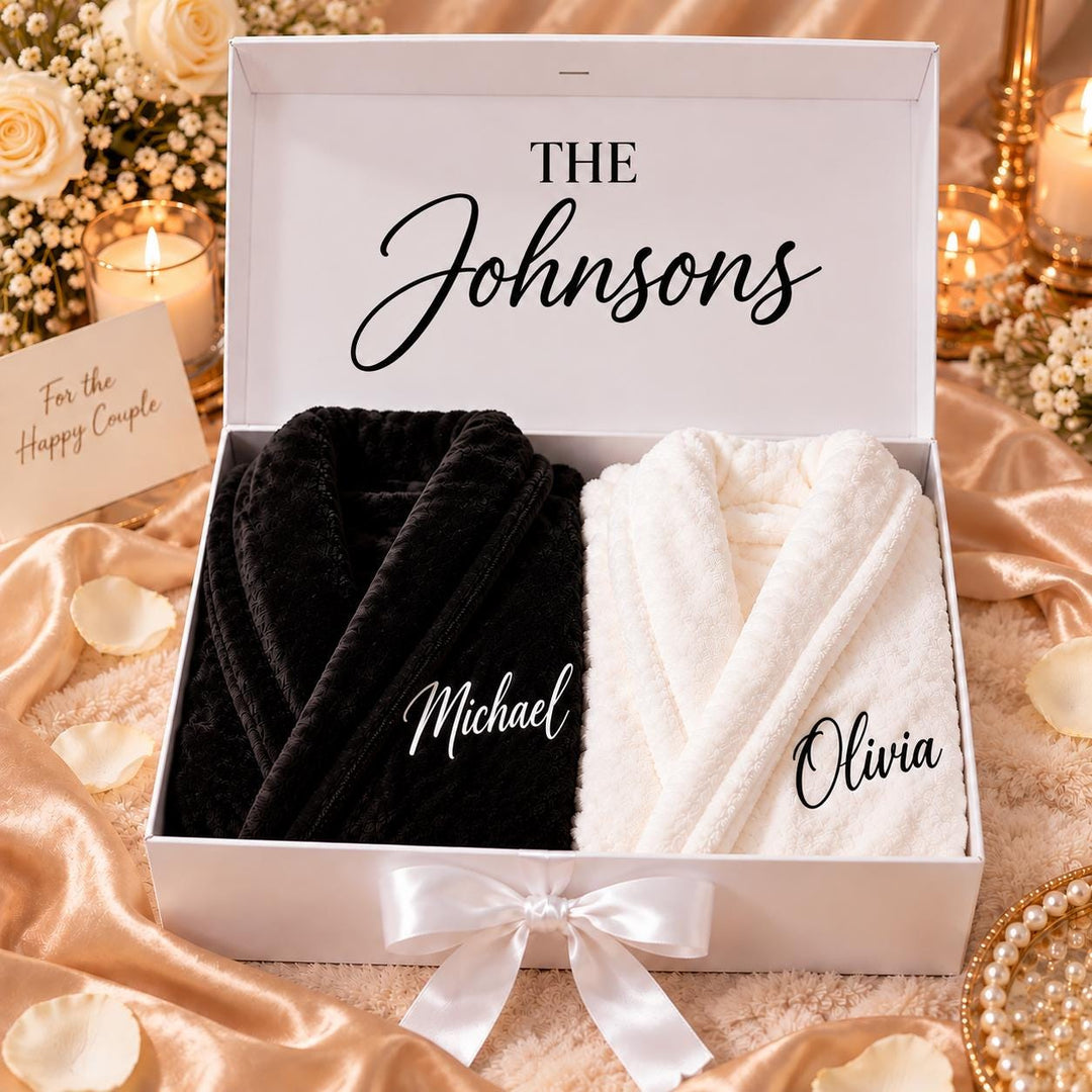 A white gift box with a bow, containing two bathrobes, one black and one white, with the names "Michael" and "Olivia" printed on them. The box is placed on a table with a floral arrangement, and there is a candle in the background.