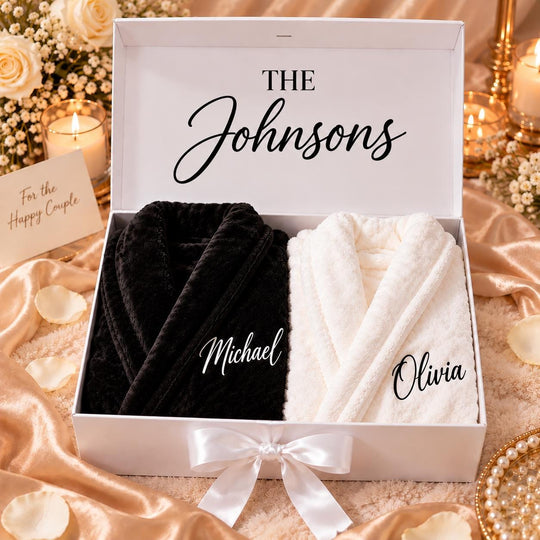 A white gift box with a bow, containing two bathrobes, one black and one white, with the names "Michael" and "Olivia" printed on them. The box is placed on a table with a floral arrangement, and there is a candle in the background.