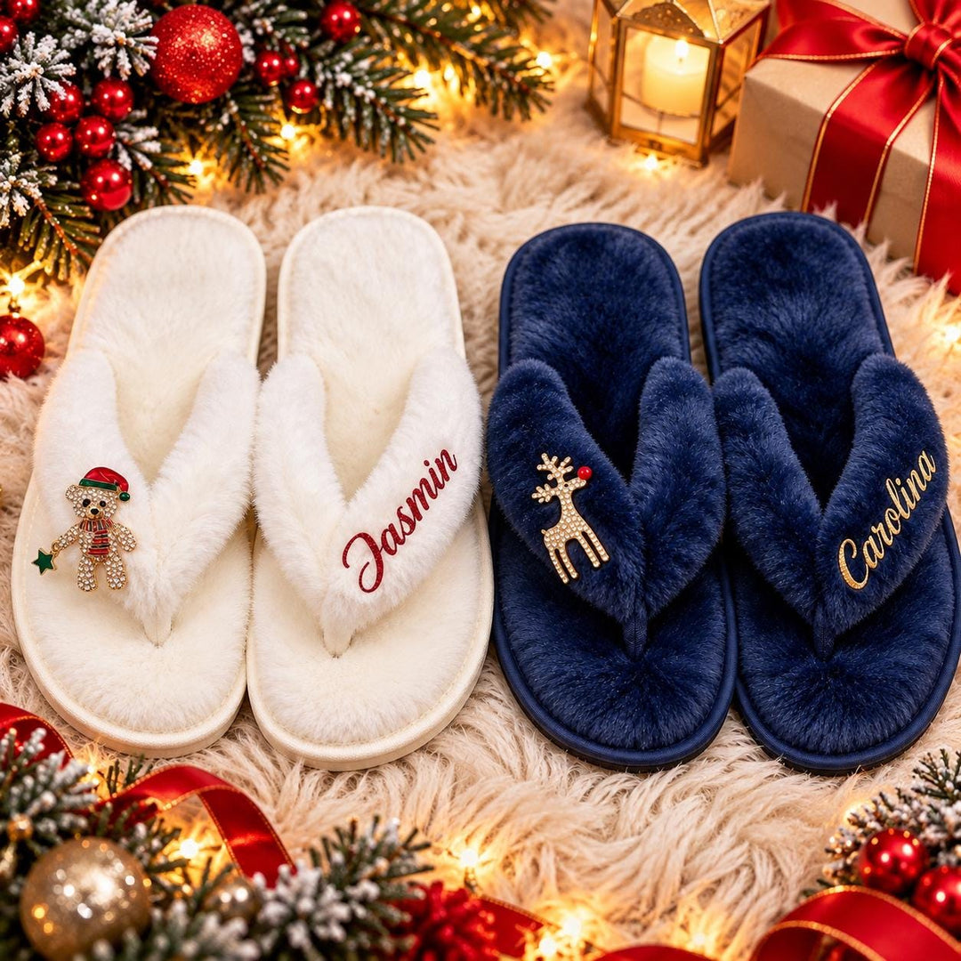 A pair of white slippers with the name "Jasmin" written on them, and a pair of blue slippers with the name "Carolina" written on them, are placed on a white shag rug with Christmas decorations and a lit lantern in the background.