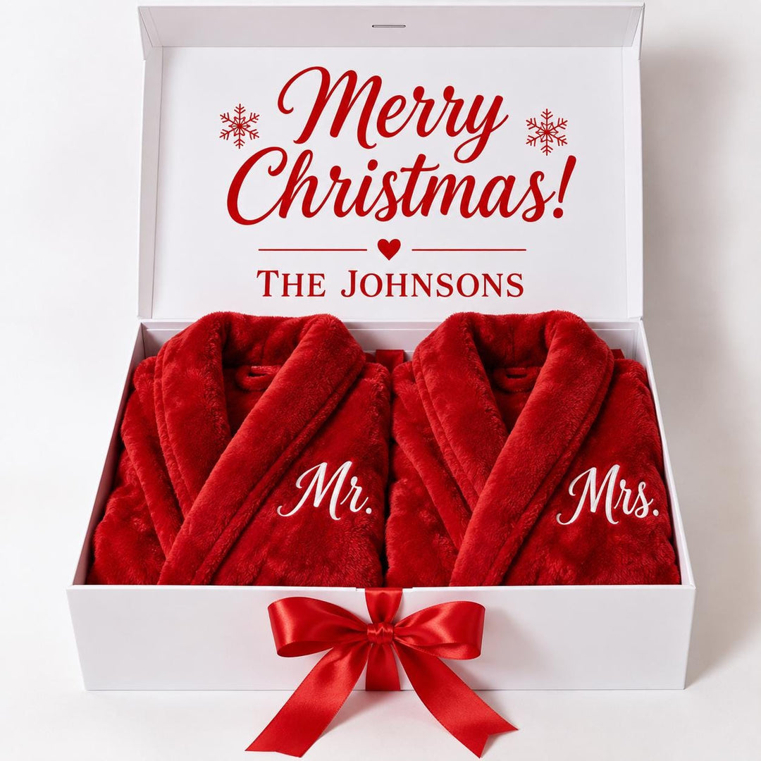 A white gift box containing two red robes with the names "Mr." and "Mrs." embroidered on them. The box is decorated with a red ribbon and snowflakes, and the text "Merry Christmas!" is displayed on the lid.